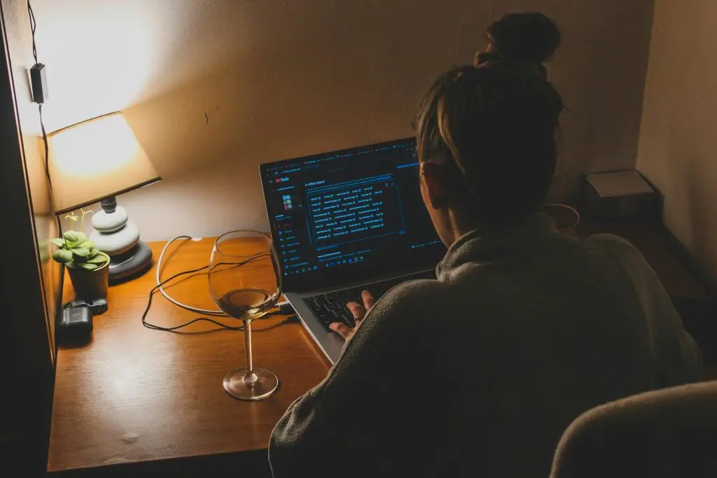 A person working from home at night, typing on a laptop with soft lamp lighting and a glass of wine.